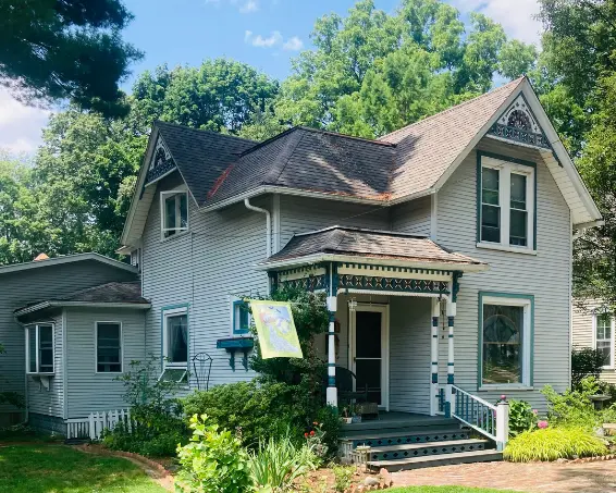 A light blue Victorian house with a dark roof and decorative trim stands among lush green trees under a bright blue sky.
